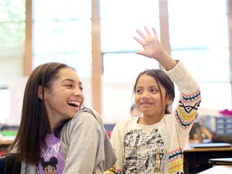 two female middle school students in classroom, one raising hand